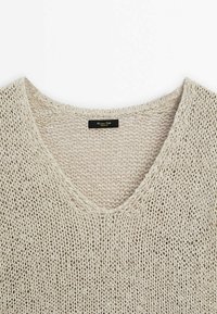 V-neck knitted sweater in light beige with an open weave texture. Label visible at the neckline. Minimalist design with soft, breathable material.