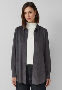 Graues Wildlederhemd mit einer glatten Textur, kragenlosem Design und langen Ärmeln. Getragen über einem weißen, gerippten Rollkragenpullover.