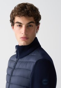 Mladý muž s kučeravými hnedými vlasmi, oblečený v námornícky modrej prešívanej bunde s pletenými rukávmi a vysokým golierom, na pozadí čistej bielej farby.