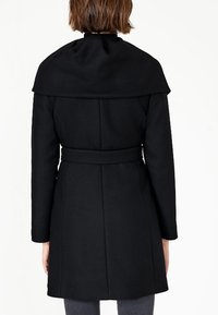 Cappotto nero realizzato in tessuto strutturato, con ampio colletto, vita con cintura, vestibilità sagomata, maniche lunghe e silhouette al ginocchio.