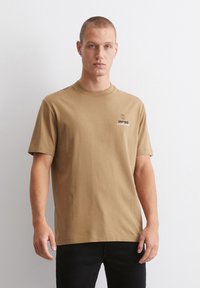 Brun bomull t-skjorte med rund hals, korte ermer og en liten globuslogo på brystet. Stoffet ser glatt og ensfarget ut.