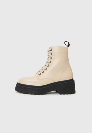 Bottines beiges avec une semelle noire épaisse et un laçage à œillets, dotées d'une languette à l'arrière et d'un logo sur le côté.