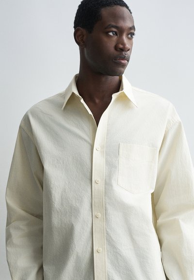 Light beige button-up shirt with a textured finish, a pointed collar, and a single chest pocket. Includes front buttons and long sleeves.