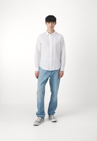 Camicia bianca a maniche lunghe con bottoni, abbinata a jeans in denim blu chiaro e sneakers con fantasia. Sfondo semplice.