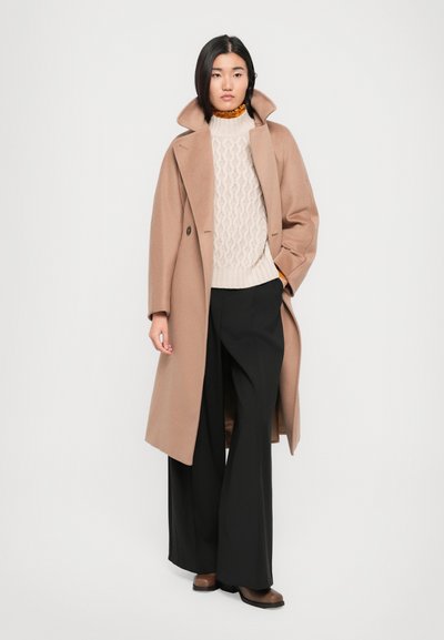 Beige wool coat with wide lapels over a cream cable-knit sweater and black wide-leg trousers. Brown shoes complete the outfit.