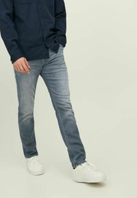 Világoskék farmernadrág, slim fit fazonban, finom kopással és ráncokkal a combokon. Fehér sportcipővel és sötétkék inget visel.