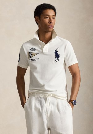 Homme portant un polo blanc avec un logo équestre bleu marine et des écussons de club nautique, associé à un pantalon blanc à cordon et une montre marron.