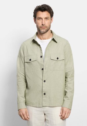 Uomo che indossa una camicia verde chiaro con bottoni e tasche con patta sopra una maglietta bianca, in piedi davanti a uno sfondo semplice.