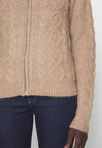 Gros plan d'une personne portant un pull zippé beige en maille torsadée et un jean bleu foncé, bras et main détendus sur le côté.