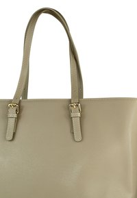 Beige draagtas gemaakt van glad synthetisch materiaal. Het heeft twee korte handvatten met gespen in goudkleur en contrasterende stiksel-details.