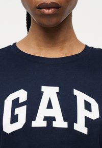 Femme portant un t-shirt bleu marine à col rond avec de grandes lettres blanches GAP sur la poitrine, ne montrant que son cou et ses lèvres.