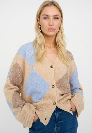 Smėlio spalvos ir mėlynos spalvos argilės raštu megztinis su priekine sagų eilėmis, banguotais rankogaliais ir minkšta tekstūra, derinamas su džinsais.