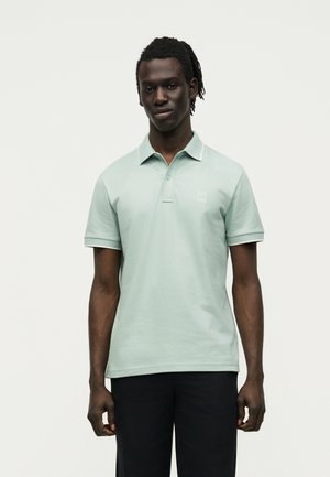 Άνδρας με κοντά dreadlocks που φοράει ανοιχτό πράσινο πόλο και μαύρο παντελόνι, στέκεται μπροστά σε ένα απλό λευκό φόντο.