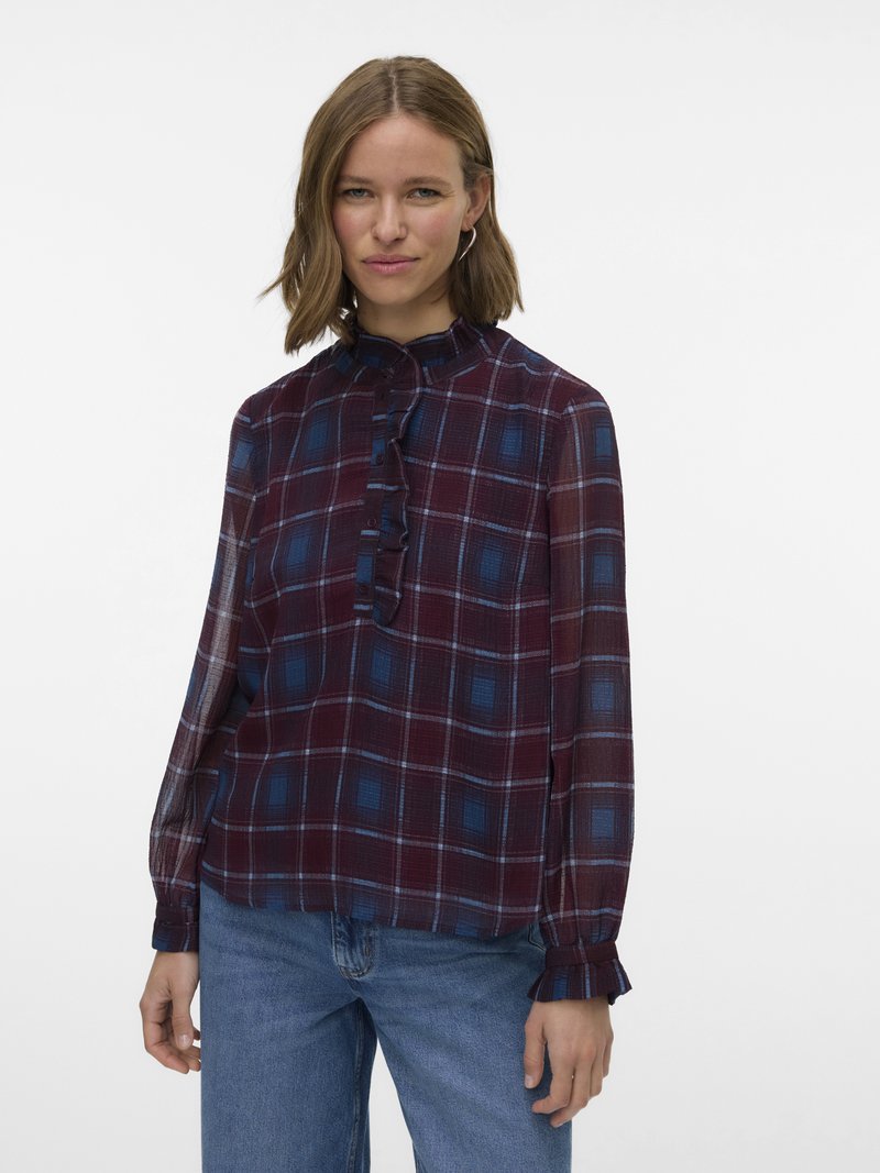 Vrouw draagt een doorschijnende bordeauxrode en blauwe geruite blouse met gerimpelde kraag en manchetten, gecombineerd met een blauwe spijkerbroek, staand tegen een witte achtergrond.