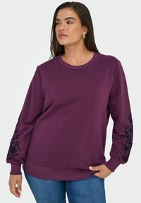 Sweatshirt violet à manches longues, avec des broderies florales noires sur les manches. Poignets et ourlet côtelés. Tissu doux et texturé.