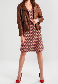 Hnědá kožená motocyklová bunda přes vzorované červené šaty s geometrickými tvary, doplněná červenými vysokými podpatky. Hladká textura, přiléhavý střih.