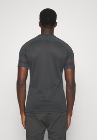 Tummanharmaa lyhythihainen paita, jossa on teksturoitu pinta ja oranssit korostukset hihoissa ja takapaneelissa.