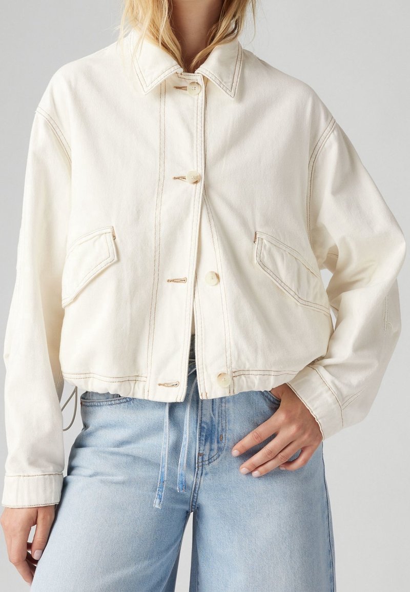 Kort vit denimjacka med knäppning framtill, spetsig krage och framfickor. Har kontrasterande beige sömmar och en avslappnad passform.