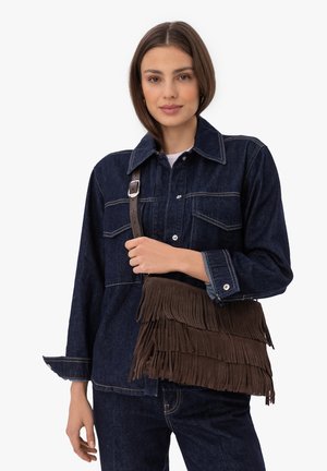 Femeie purtând jachetă și blugi din denim închis, ținând o geantă crossbody maro din piele întoarsă cu franjuri și curea cu cataramă.