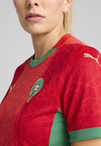 Röd och grön sporttröja med geometriska mönster, rund halsringning och en gul Puma-logga på axeln. Marockos emblem visas.