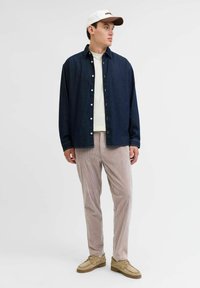 Denimskjorte med knaplukning foran i mørkeblåt, parret med beige fløjl-bukser. Modellen bærer en hvid kasket med brune detaljer.