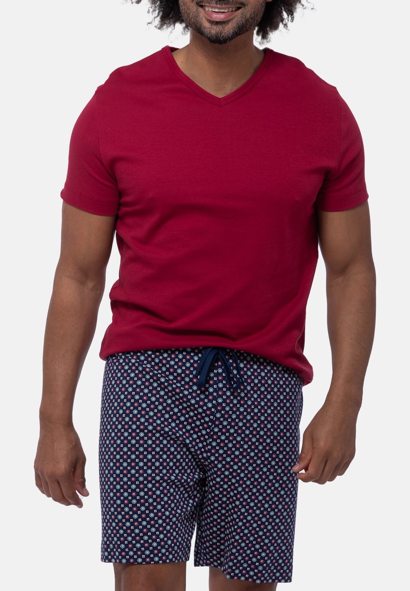 Homme portant un t-shirt à manches courtes rouge foncé avec un col en V et un short bleu marine à petit motif floral blanc et rouge sur un fond uni.