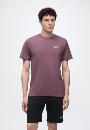 Homme aux cheveux bouclés portant un t-shirt violet Puma et un short noir Puma, debout devant un fond blanc uni.