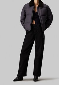 Osoba ubrana w ciemną kurtkę puchową z kołnierzem z owczej wełny, czarny krótki top zapinany na guziki, szerokie czarne dżinsy oraz czarne botki na obcasie.