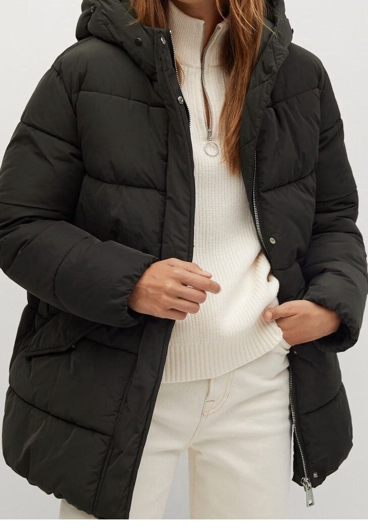 Femme portant une doudoune noire par-dessus un pull en maille blanc avec un col zippé et un pantalon clair, mains ajustant les poches de la veste.