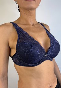Sostén de encaje azul marino con diseño de escote en pronunciación, patrón floral, tirantes ajustables y un charm en forma de corazón en el centro, con una base de tela suave.