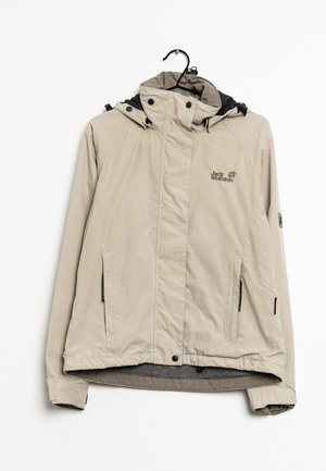 Veste de plein air beige clair avec capuche, poches zippées et logo Jack Wolfskin sur la poitrine, suspendue sur un cintre noir devant un fond blanc.
