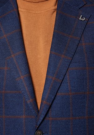 Niebieski materiał w kratę na marynarkę z brązowymi akcentami, noszony na pomarańczowym golfie. Zauważalne szczegóły to małe metalowe logo na klapie marynarki.