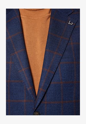 Niebieski materiał w kratę na marynarkę z brązowymi akcentami, noszony na pomarańczowym golfie. Zauważalne szczegóły to małe metalowe logo na klapie marynarki.