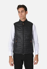 Gilet imbottito nero con colletto alto, zip frontale e due tasche con zip accentuate da tiranti arancioni. Indossato sopra una camicia bianca.
