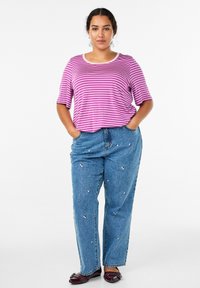 Naine seisab käed taskus, seljas lillat ja valget triibulist särki, broderi täis sinised teksad ja tumepunased korterid.