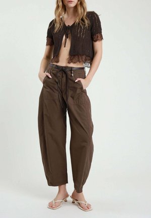Donna che indossa un cardigan nero corto con bordi in pizzo, pantaloni larghi marroni con coulisse e sandali beige con tacco, in piedi con le mani nelle tasche.