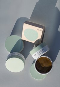 Barattoli di un verde chiaro opaco con coperchi rotondi e etichette bianche, contenenti un gel marrone; accompagnati da una scatola beige con un design circolare teal.