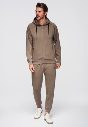 Homme portant un sweat à capuche zippé marron assorti et un pantalon de jogging avec des cordons noirs et des baskets blanches, debout devant un fond uni.