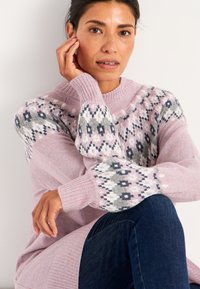 Rosa gestrickter Pullover mit einem geometrischen Muster in Grau, Blau und Weiß an den Schultern und langen Ärmeln, kombiniert mit dunklen Jeans.