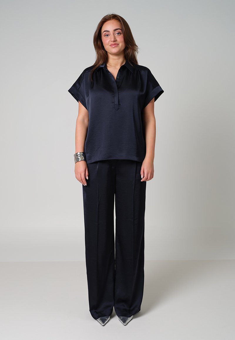 Marineblauwe zijde blouse met een knoopsluiting en korte, brede mouwen, gecombineerd met bijpassende high-waisted, brede broekspijpen. Zilveren enkellaarsjes.