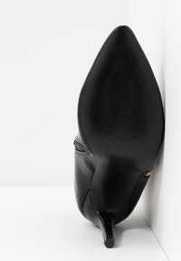 Scarpa nera con punta a stiletto posata sul tallone contro un muro e un pavimento bianchi.