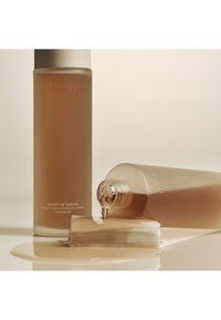 Huxley Secret of Sahara Conditioning Essence Toner in een frosted glazen fles; de vloeistof is goudbeige en morst op een lichtgekleurde ondergrond.