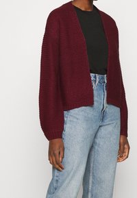 Burgundyröd stickad cardigan med en avslappnad passform, öppen front och ribbad ärm, matchad med högmidjade ljusblå jeans. Slät textur.