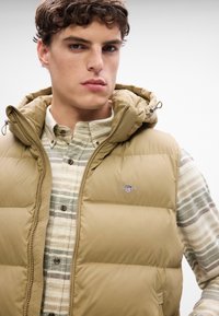 Gilet imbottito khaki con cappuccio, dotato di un logo sul petto, indossato sopra una camicia di flanella a righe verdi e beige con chiusura a bottoni.