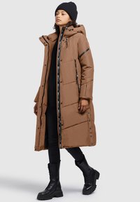 Brauner Puffer-Mantel mit Kapuze, ausgestattet mit einem Reißverschluss, Seitentaschen und schwarzen Akzenten. Kombiniert mit schwarzen Leggings und klobigen Schnürstiefeln.
