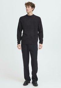 Pulover negru tricotat cu guler rotund, asortat cu pantaloni închiși la culoare și pantofi negri. Textura puloverului pare moale și călduroasă.