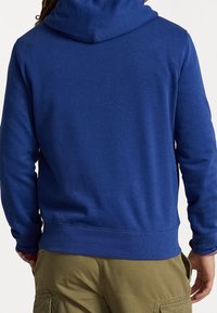 Man i blå huvtröja och olivgröna cargobyxor sedd bakifrån med händerna vid sidorna.