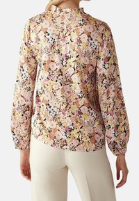 Kwiatowa bluzka w pastelowych kolorach z długimi rękawami i marszczonymi mankietami. Posiada wysoki kołnierz oraz skomplikowane wzory kwiatowe na jasnym tle.