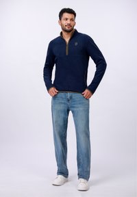 Pull-over en polaire bleu marine avec un zip quart, un logo sur la poitrine et des accents vert olive. Porté avec un jean bleu clair et des baskets blanches.