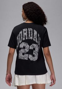 Juodas marškinėliai su dideliu „JORDAN“ ir „23“ raštu, atspausdintu nėriniuotomis linijomis ant nugaros. Trumpomis rankovėmis ir laisvu siluetu. Švelni medvilnės tekstūra.
